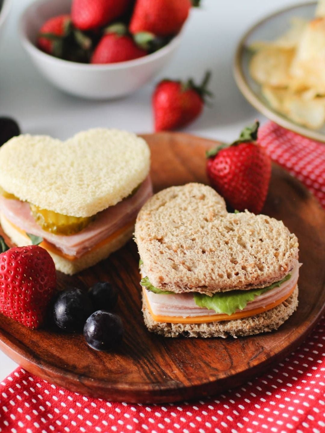 Dos sándwiches en forma de corazón con jamón, queso, lechuga y pepinillo en un plato de madera, rodeados de fresas y uvas
