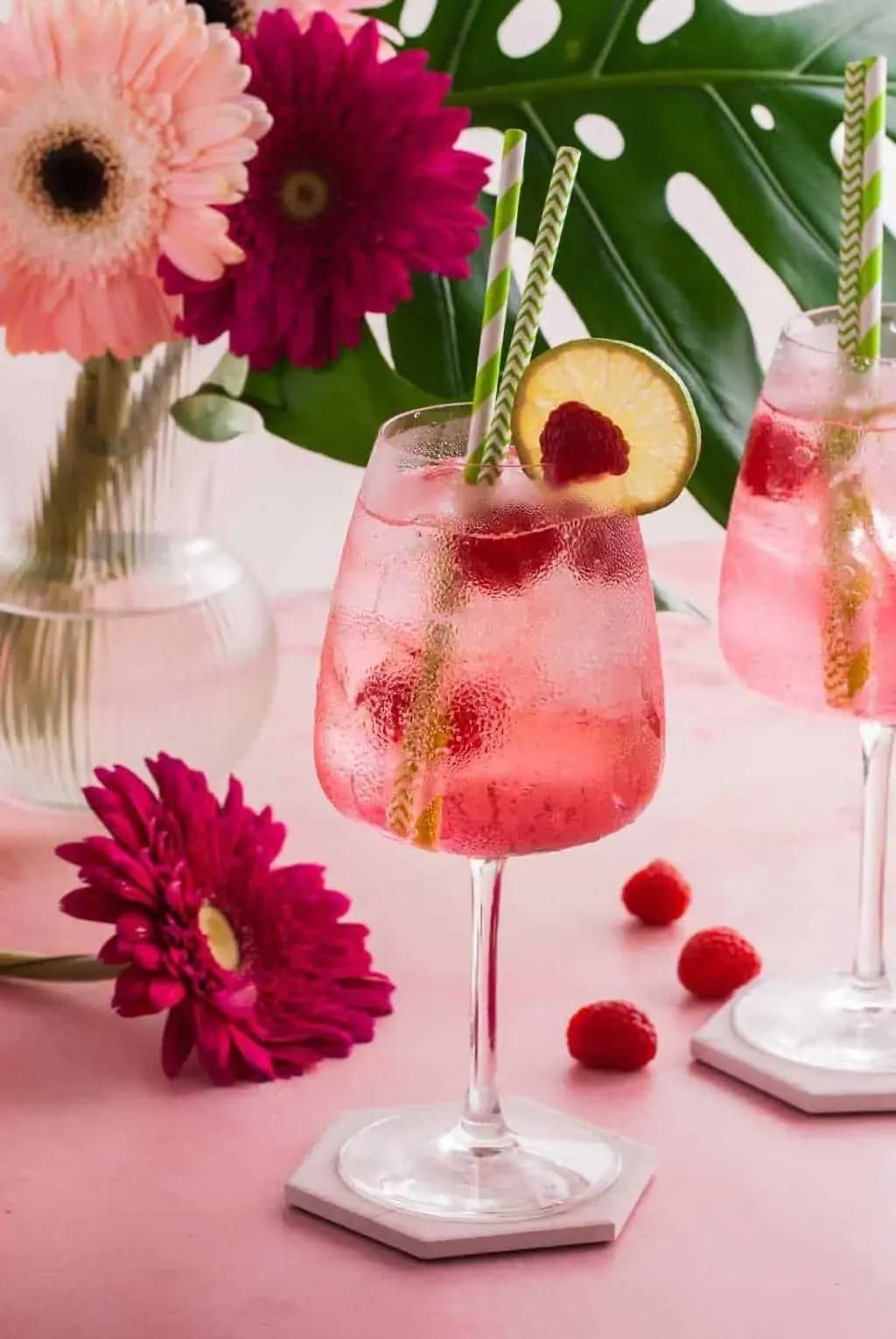 Dos copas con cócteles rosados, cada una con una rodaja de limón y una frambuesa. Fondo con flores y hojas decorativas