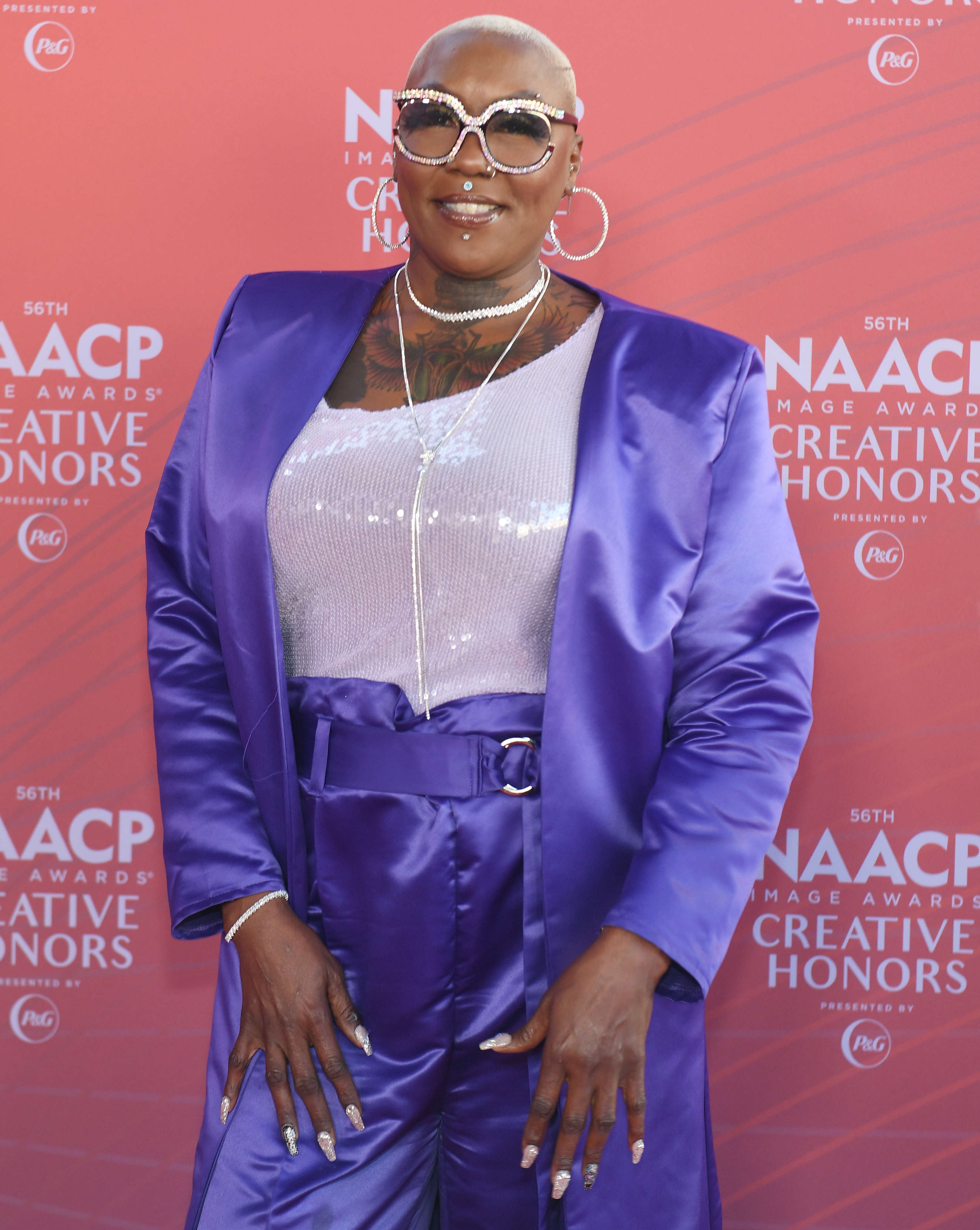 Person i elegant satengdress og glitrende topp på NAACP Image Awards. De har kort hår og bruker solbriller og lagdelte halskjeder