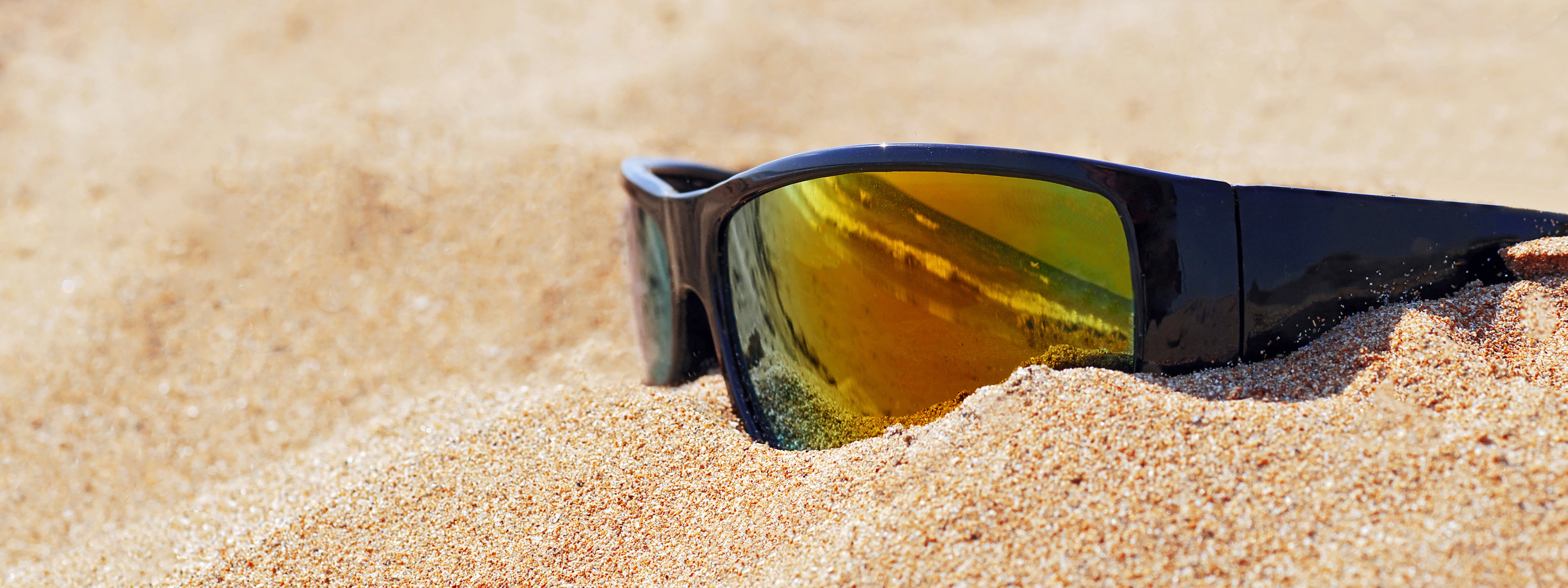 Sunglasses partially buried in sand on a beach, reflecting a clear sky