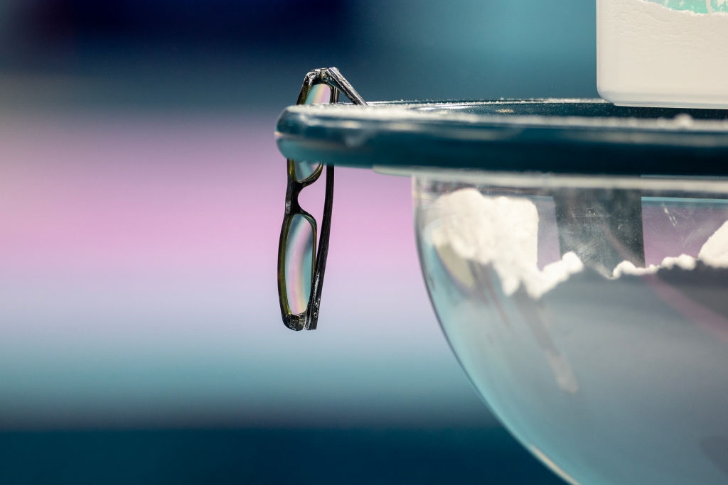 A brace  of eyeglasses rests connected  the borderline   of a vessel  filled with powder, against a blurred background