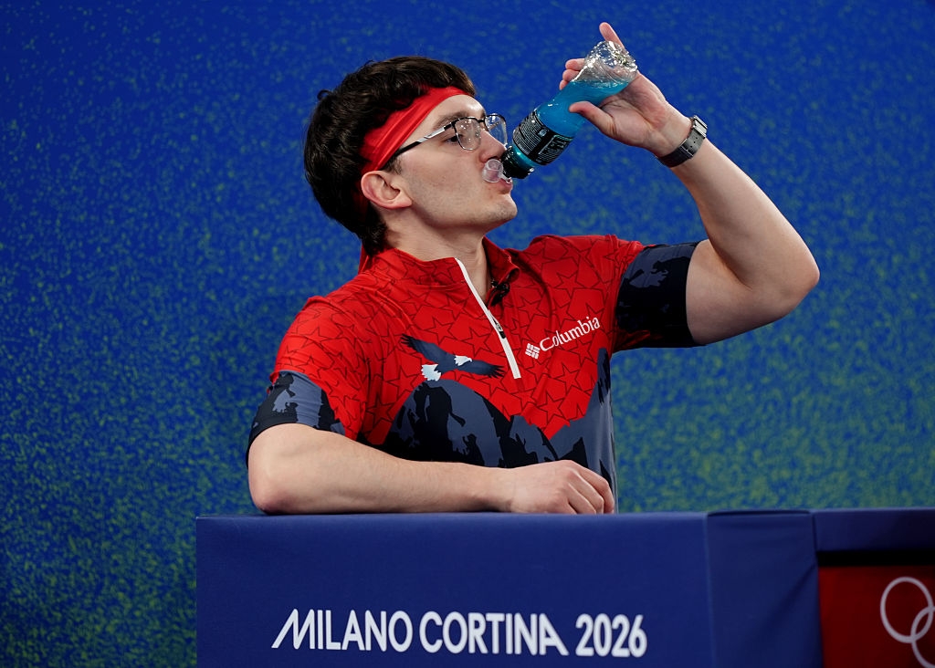 Person successful  sportswear with a headband drinks a bluish  beverage astatine  an diversion  event. "Milano Cortina 2026" is disposable   successful  the background