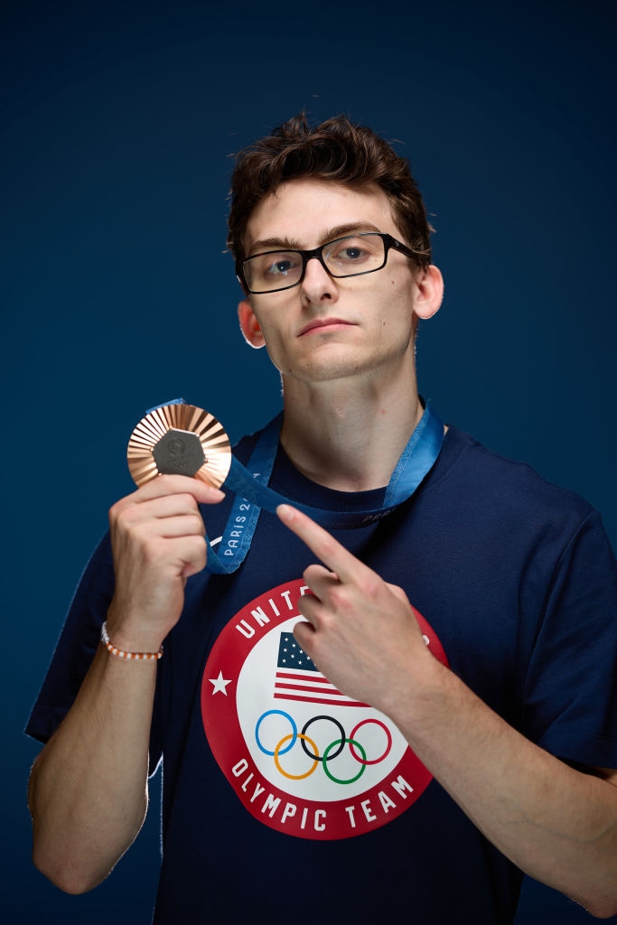 Person successful  glasses with a superior   look  holds and points to a bronze medal with "Paris" visible, wearing a U.S. Olympic Team t-shirt