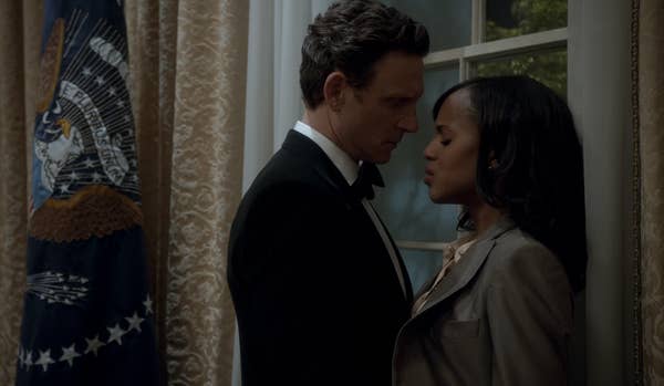 A couple stands closely in an elegant room next to an emblem flag, sharing an intense moment. Both in formal attire, with the woman in a blazer