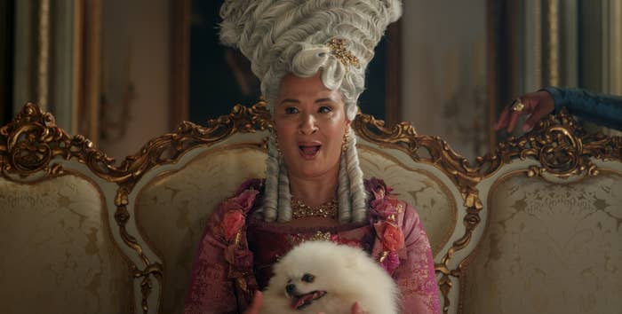 A person in elaborate historical attire, including a tall, ornate wig, sits on an ornate couch holding a fluffy dog, exuding elegance and grandeur