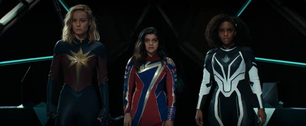Three people stand in superhero costumes on a set with dark lighting, each featuring unique designs and emblems