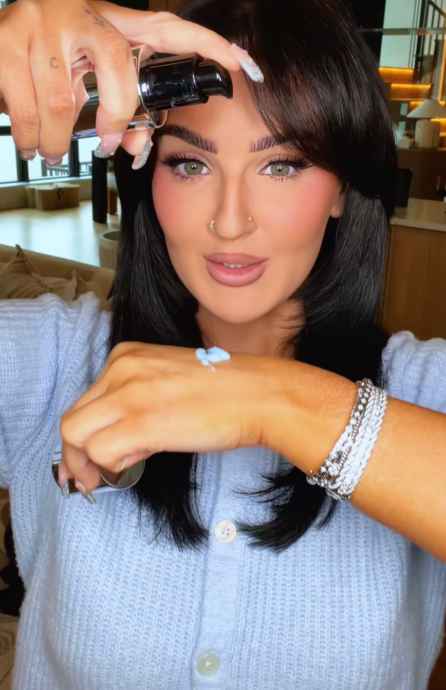 Person applying skincare merchandise  to backmost  of hand, wearing a button-up apical  and stacked bracelets, with a focused expression