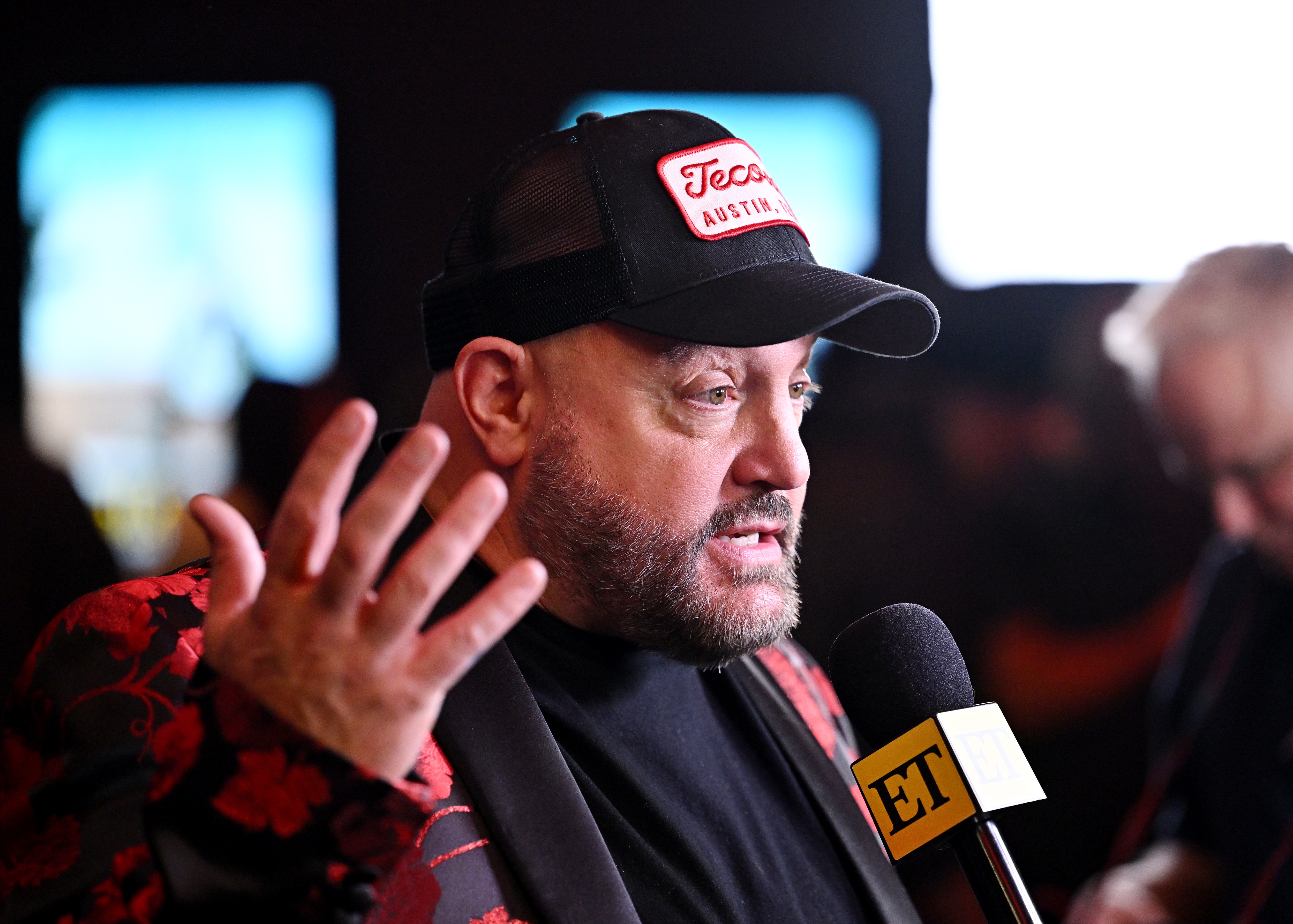 A antheral   wearing a headdress  and a floral-patterned overgarment   speaks into an ET microphone astatine  an event