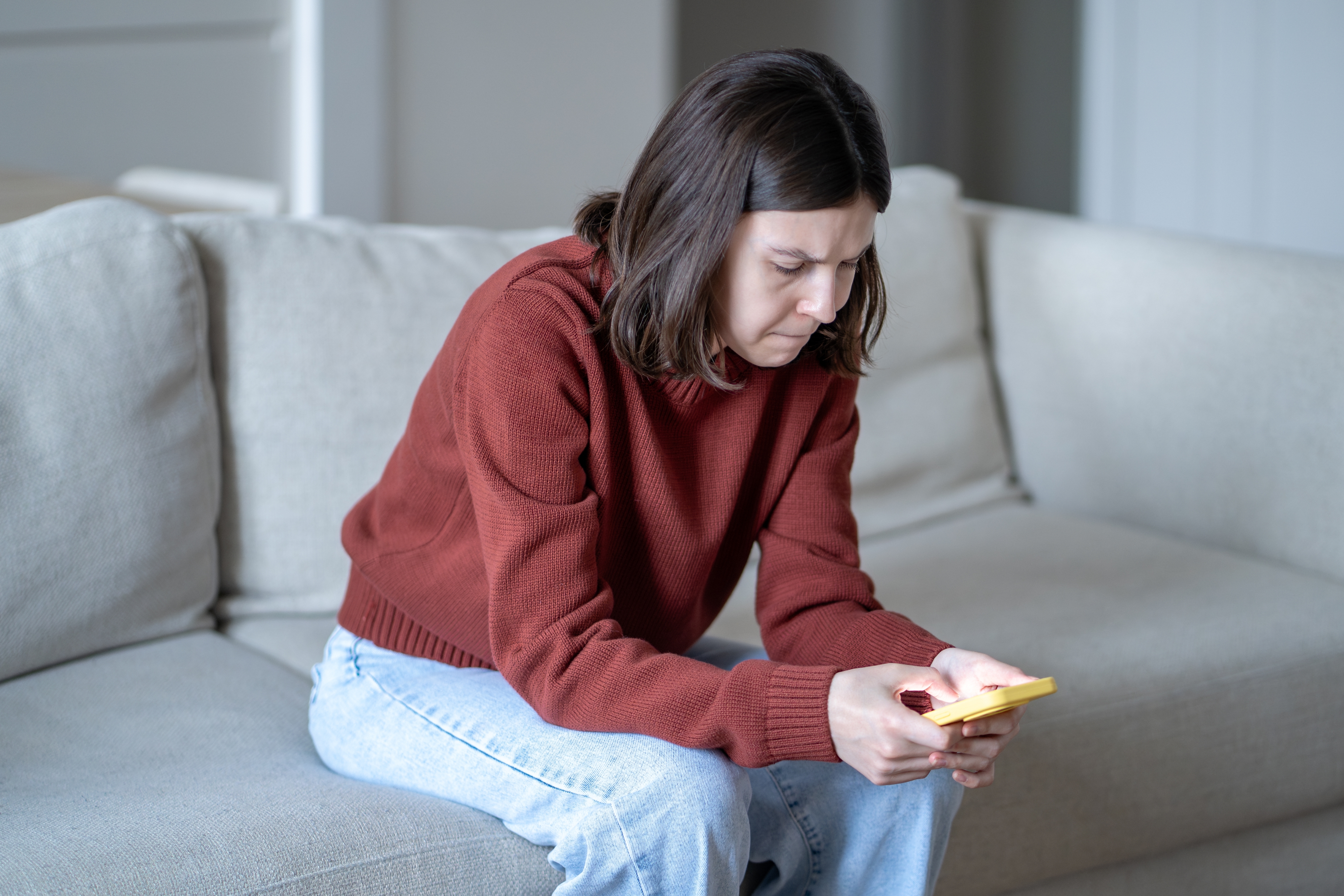 Person sitting connected a sofa, looking astatine a smartphone with a focused expression, wearing a casual sweater and jeans