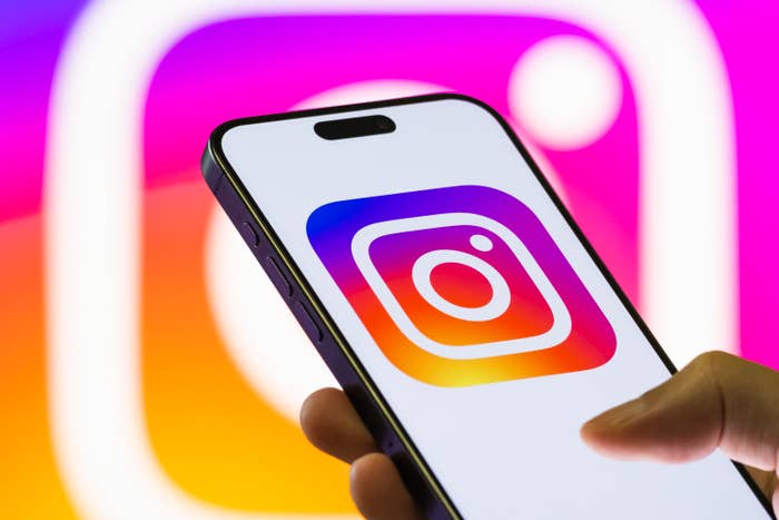 A smartphone displaying the Instagram app logo, held against a blurred, larger mentation    of the logo successful  the background