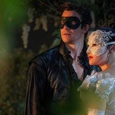 Pareja en un baile de máscaras; él lleva traje de cuero y antifaz negro, ella viste un elegante vestido con antifaz blanco