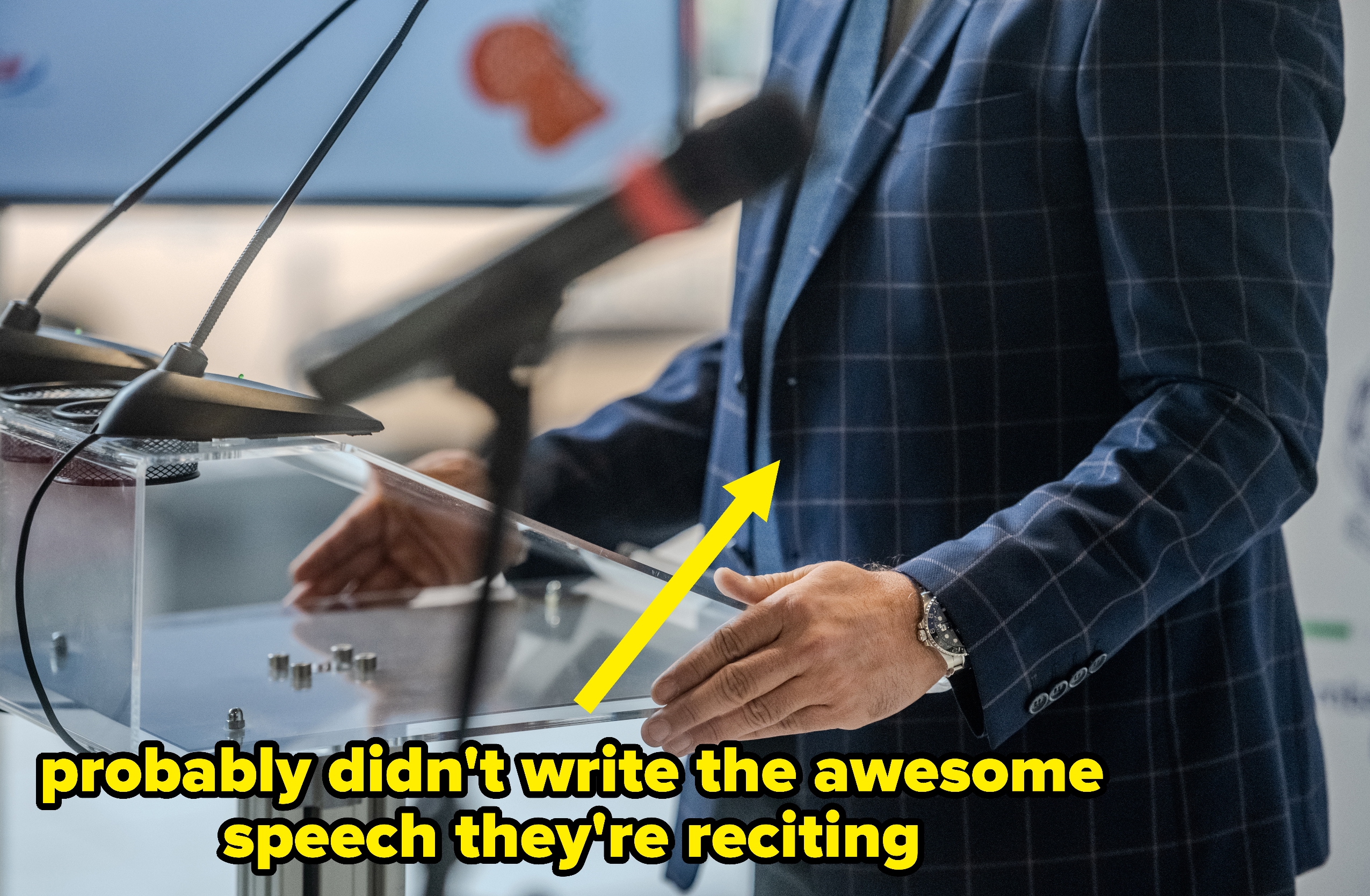 Person i rutete dress snakker ved et tydelig podium med en mikrofon, gestikulerer med hendene mens han henvender seg til et publikum