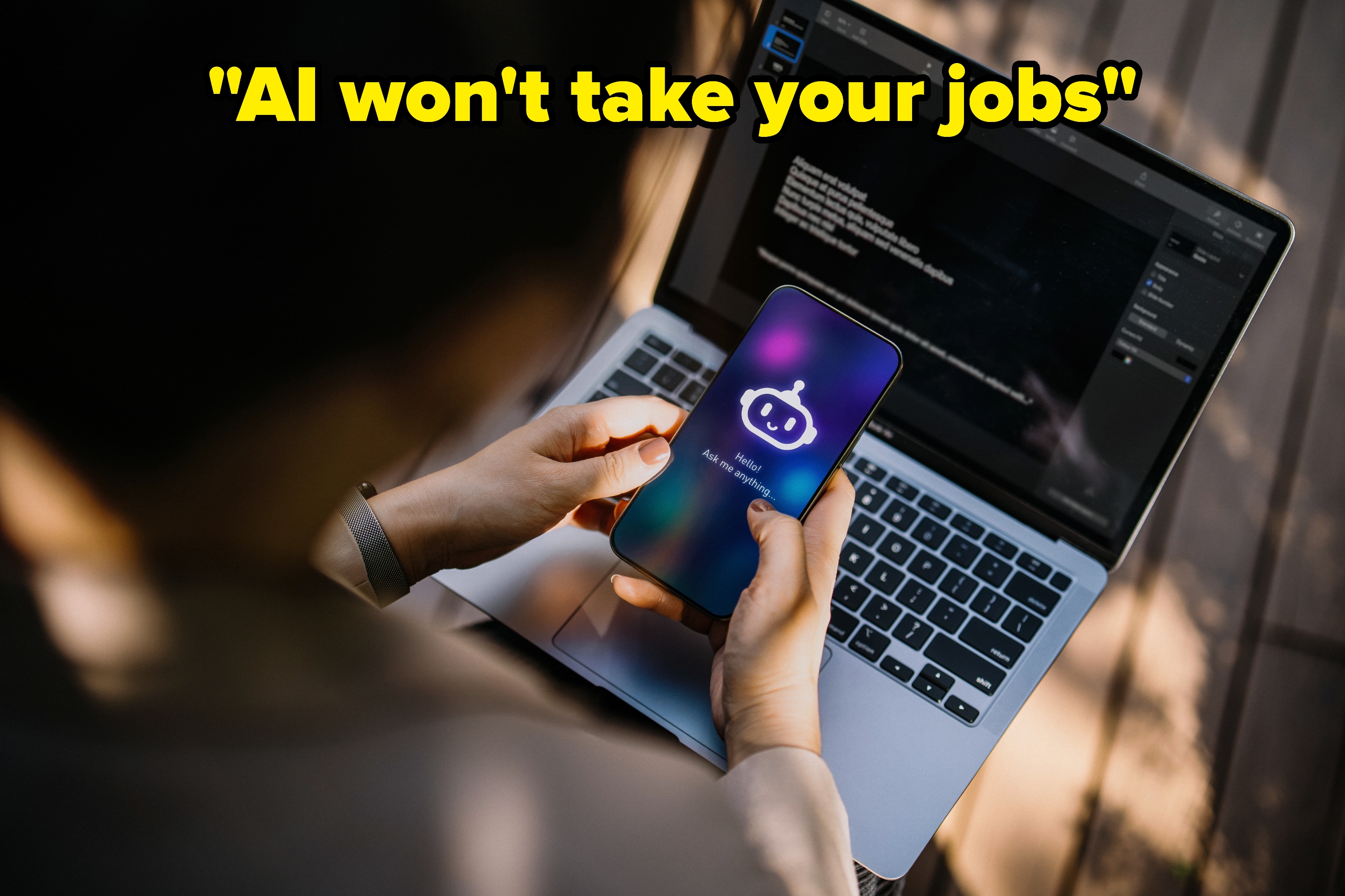 Person som holder en smarttelefon som viser et robotikon og tekst, mens han bruker en bærbar datamaskin med kode på skjermen