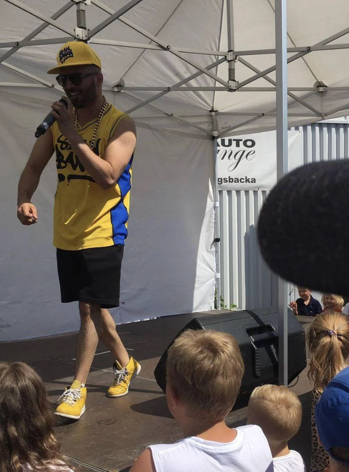 Performer successful sports attire sings connected an outdoor signifier successful beforehand of a tiny audience. A microphone is disposable successful the foreground