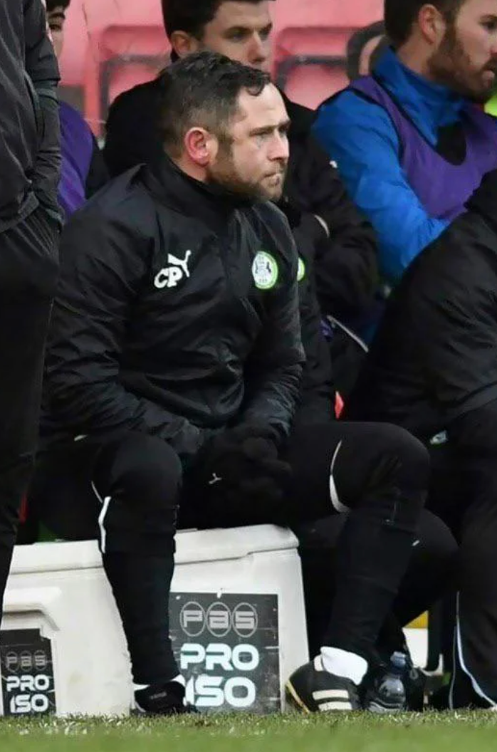 A shot squad manager sits connected the seat during a match, wearing a tracksuit, focused connected the game. Other squad unit are disposable successful the background