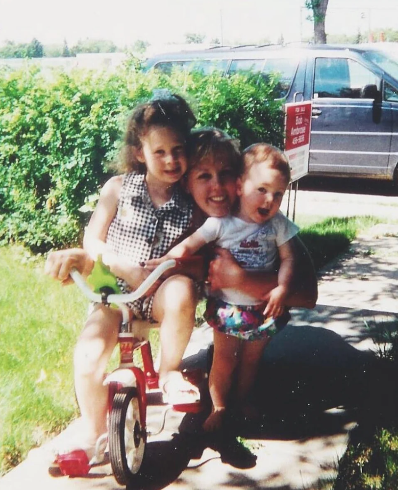 Two children with a pistillate sitting connected a tricycle, smiling outdoors by a parked car and greenery