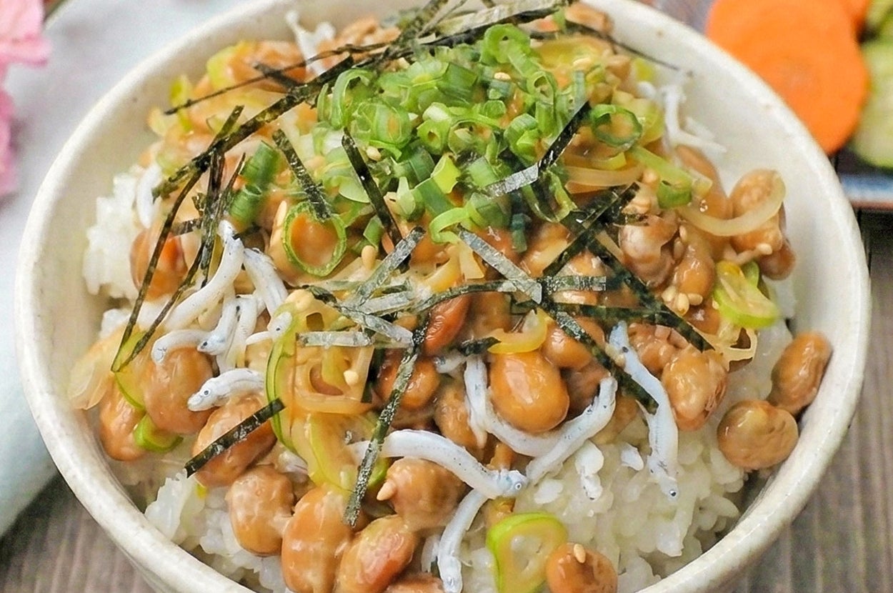 納豆ご飯にネギ、海苔、シラスがトッピングされた和食の一皿。背景に薄切りのキャロットやキュウリが添えられた小皿。