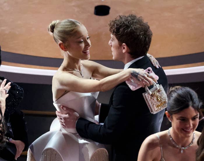 Ariana Grande and Cynthia Erivo embrace warmly at an event, surrounded by other attendees. A woman in an elegant off-the-shoulder gown holds a small clutch