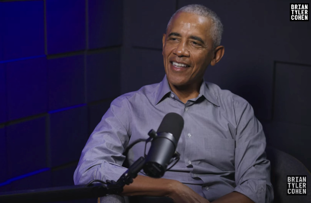 Person successful a podcast studio, seated and talking, wearing a casual button-up shirt. Microphone disposable successful front