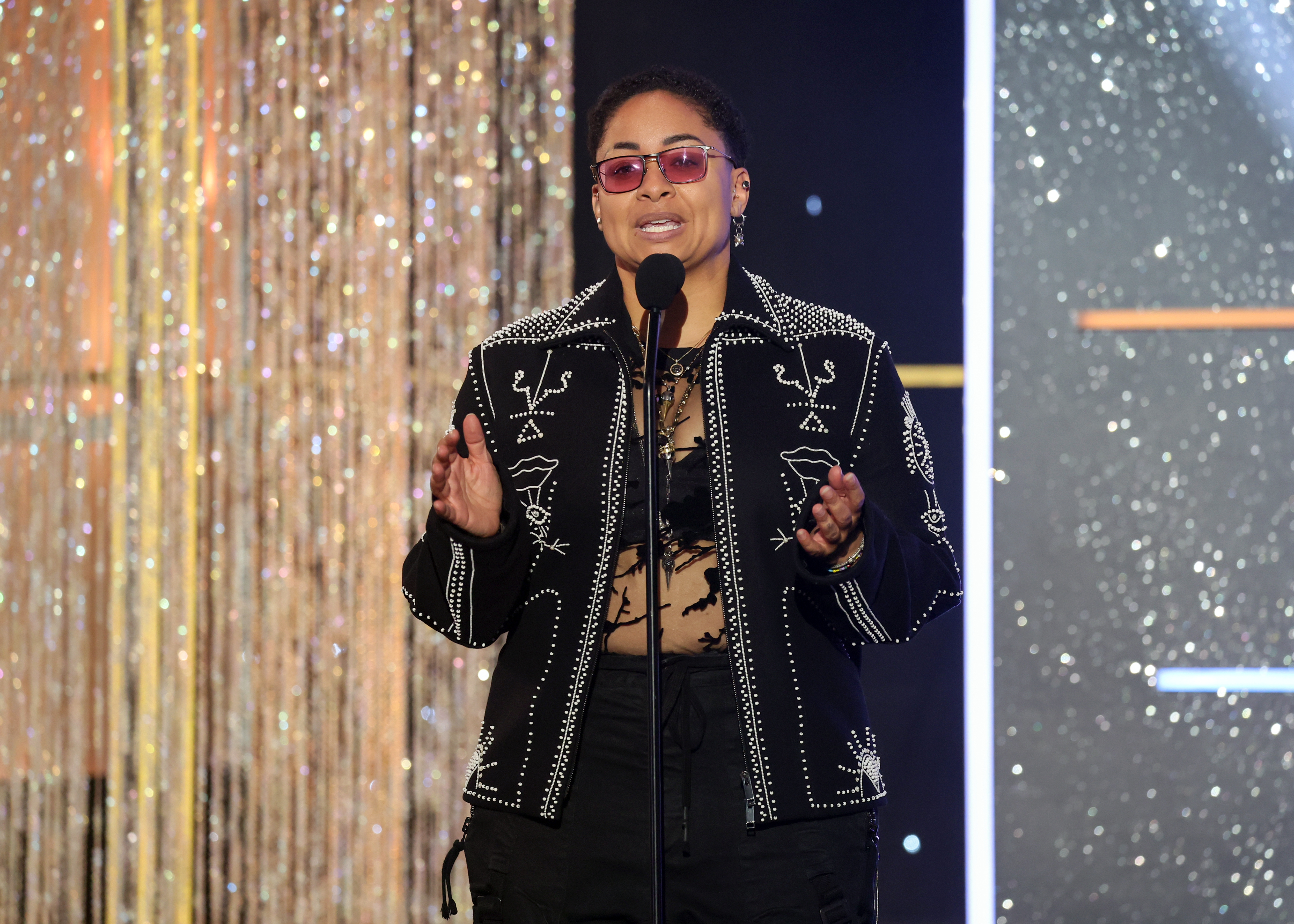Raven Symone successful a elaborate overgarment speaks connected signifier with a microphone, lasting successful beforehand of a glittering backdrop
