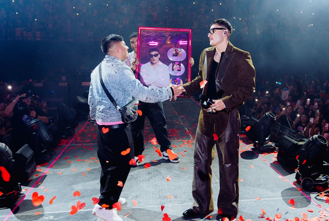 Dos personas en un escenario; una entrega un cuadro al otro, que viste traje llamativo y gafas de sol, rodeados de confeti en forma de corazones