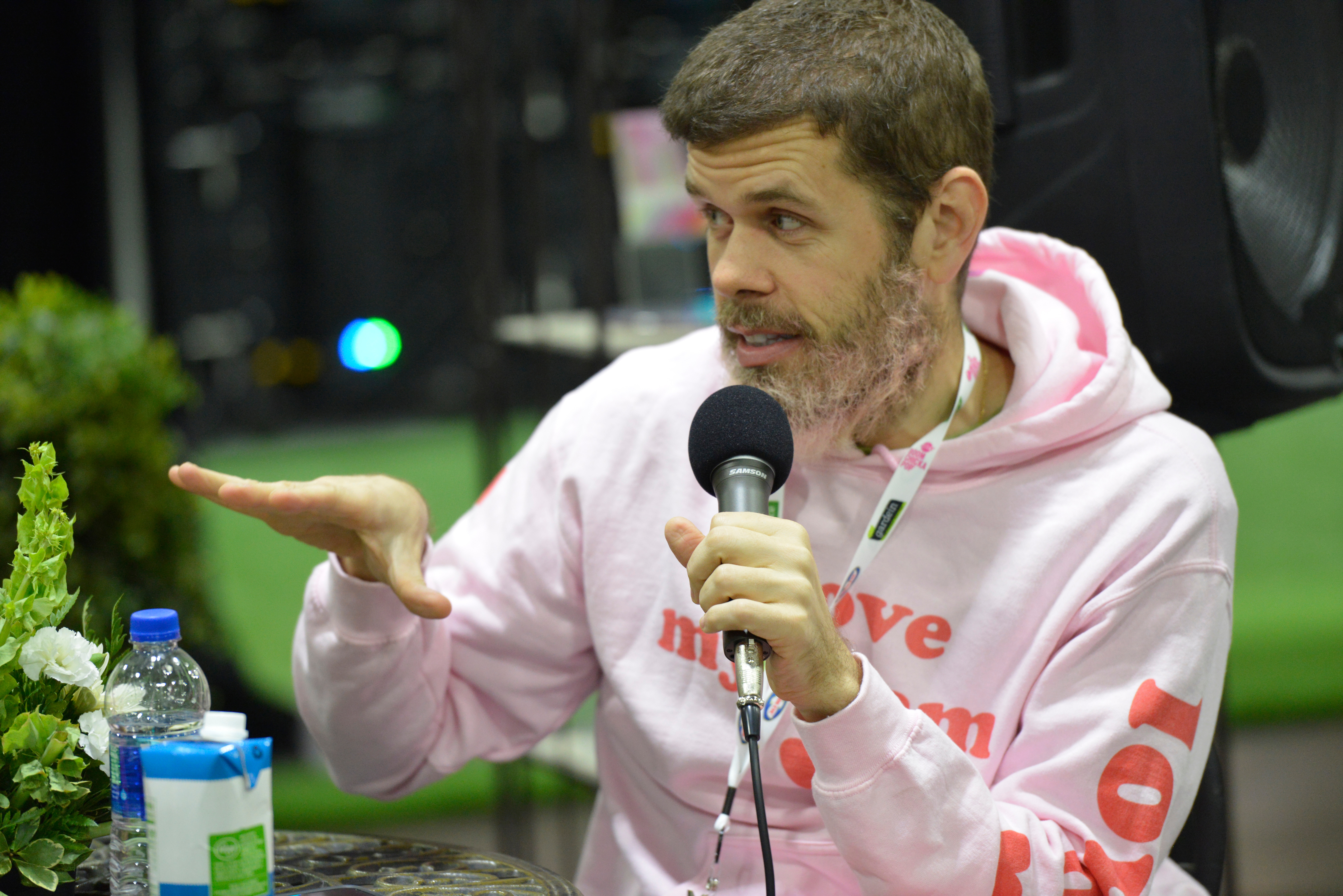 Perez Hilton successful a casual hoodie speaks into a microphone astatine an indoor event, gesturing with his hand