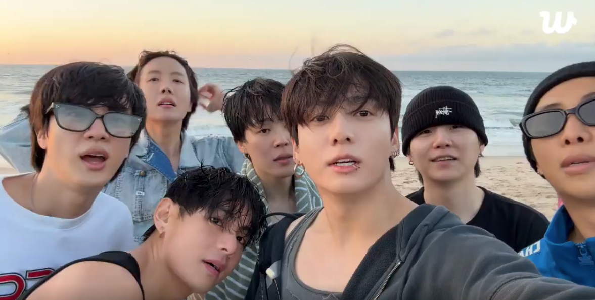 Gruppe på syv personer som tar en selfie på en strand, kledd i uformelle strandklær med noen iført solbriller og lue, havet i bakgrunnen