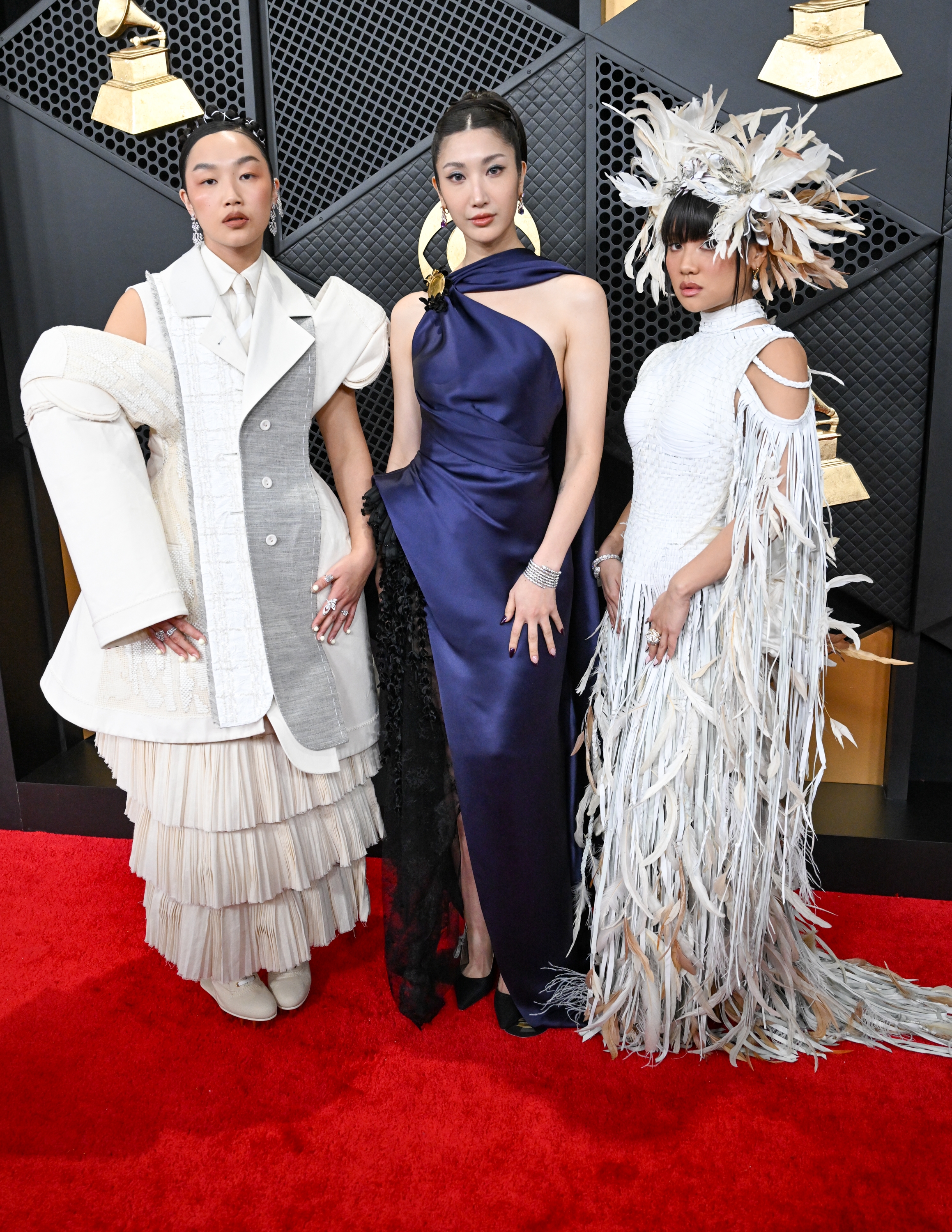 Tre individer står på en rød løper. En går i lagdelt antrekk, en annen i en elegant kjole, og den tredje i en fjærkledd kjole med hodeplagg