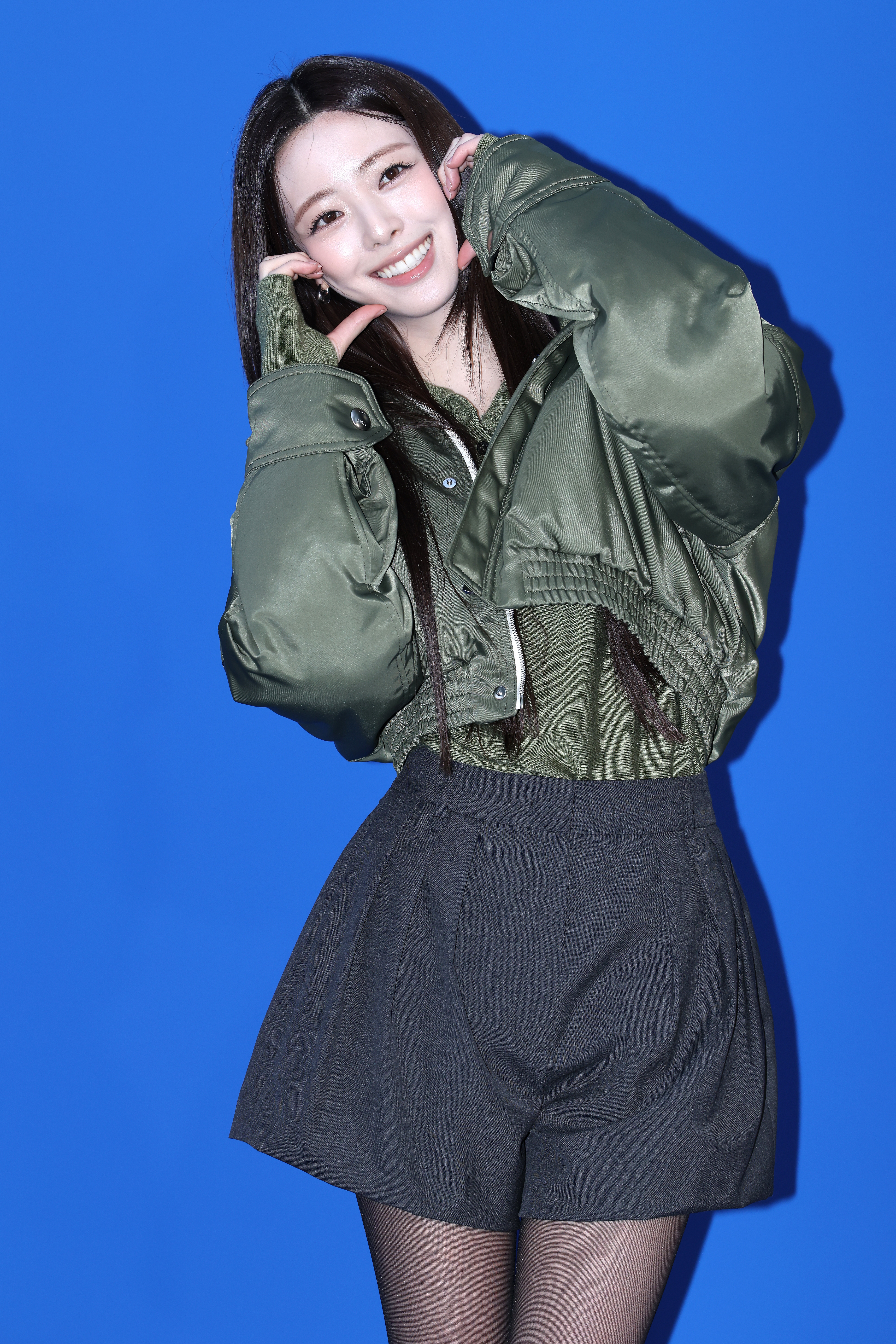 Person som poserer lekent mot et blått bakteppe, iført en grønn pufferjakke og grå shorts, smiler med hendene som rammer inn ansiktet sitt