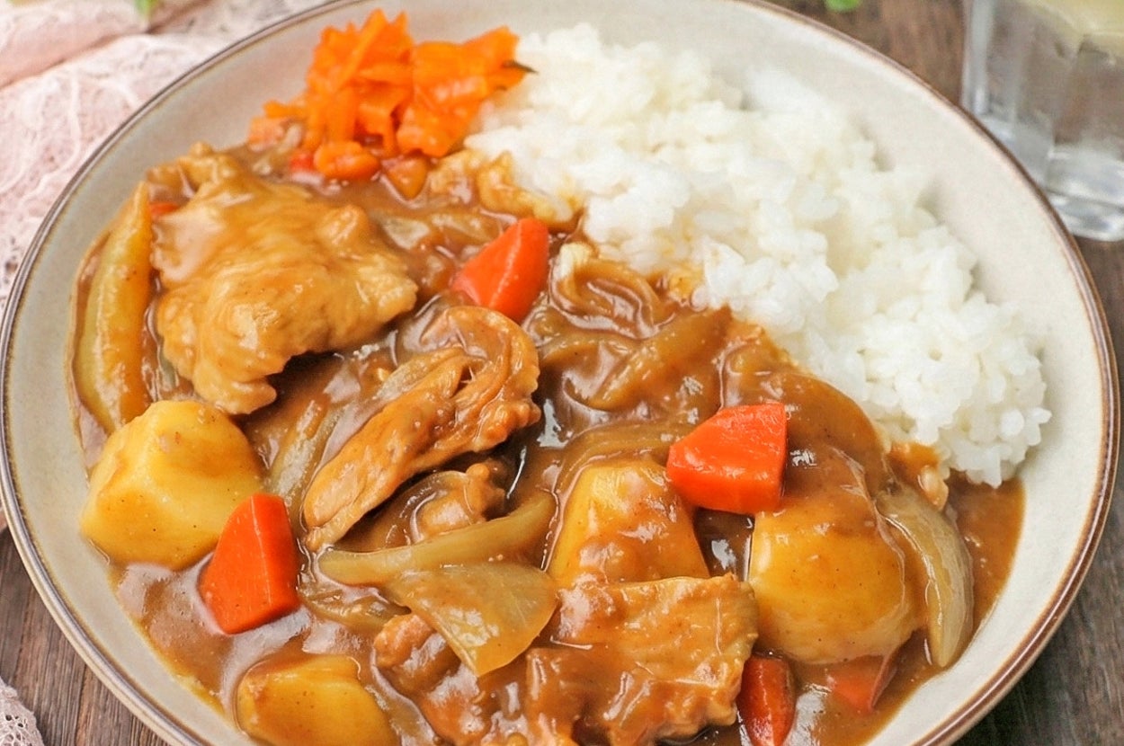 カレーライスの皿、具材に鶏肉、じゃがいも、にんじん、玉ねぎ。横にレモン入りの水。背景に植物。