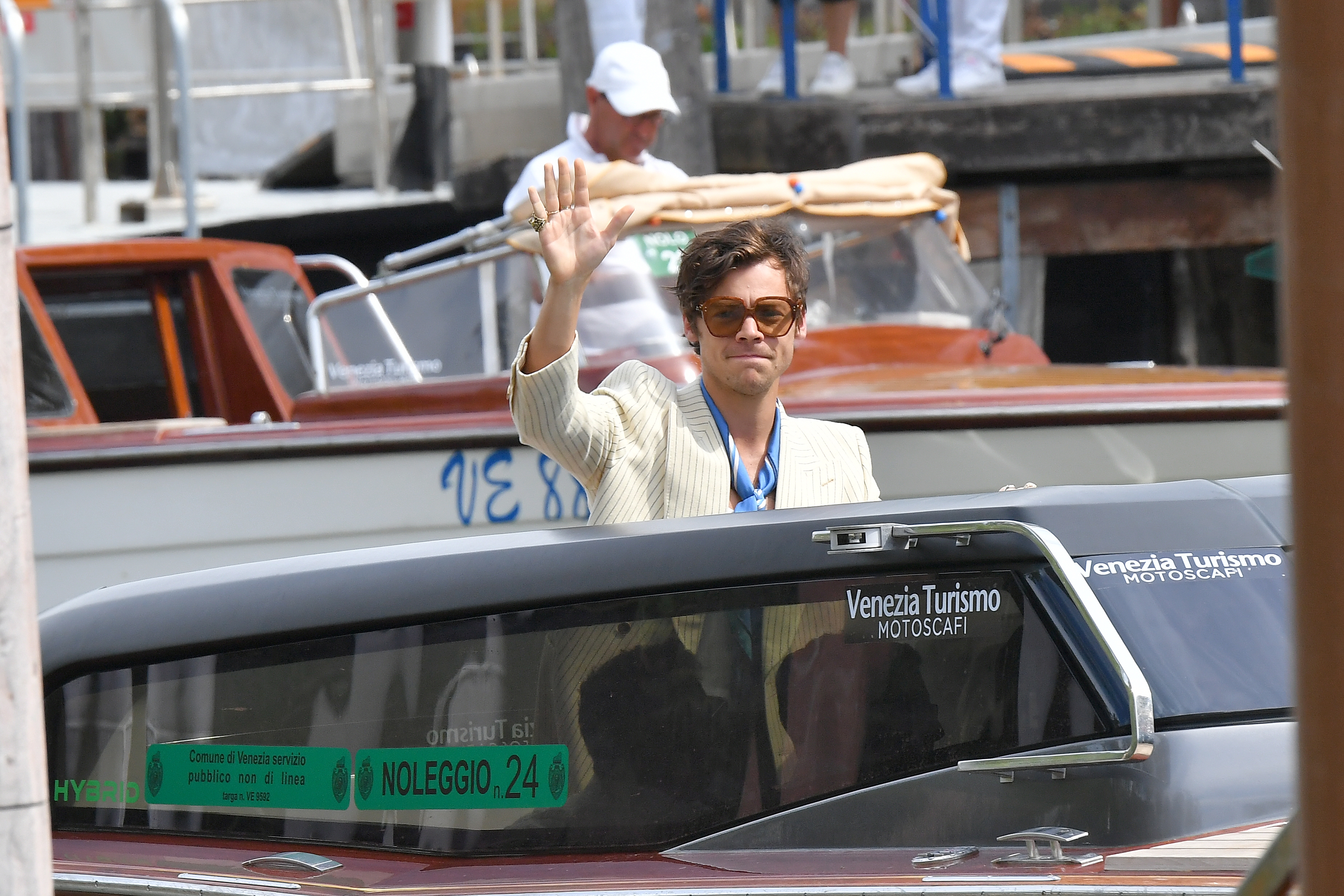Person in a light blazer waves from a boat at a public event, creating an approachable and casual style