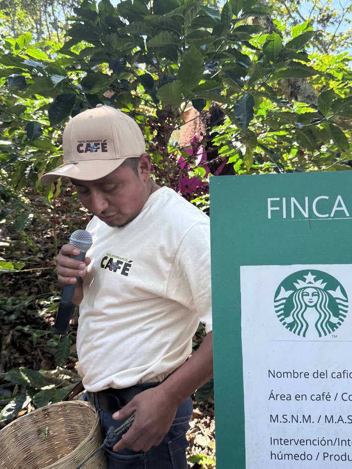 Hombre con camiseta y gorra de "Café" sostiene un micrófono junto a un cartel de Starbucks en una plantación de café