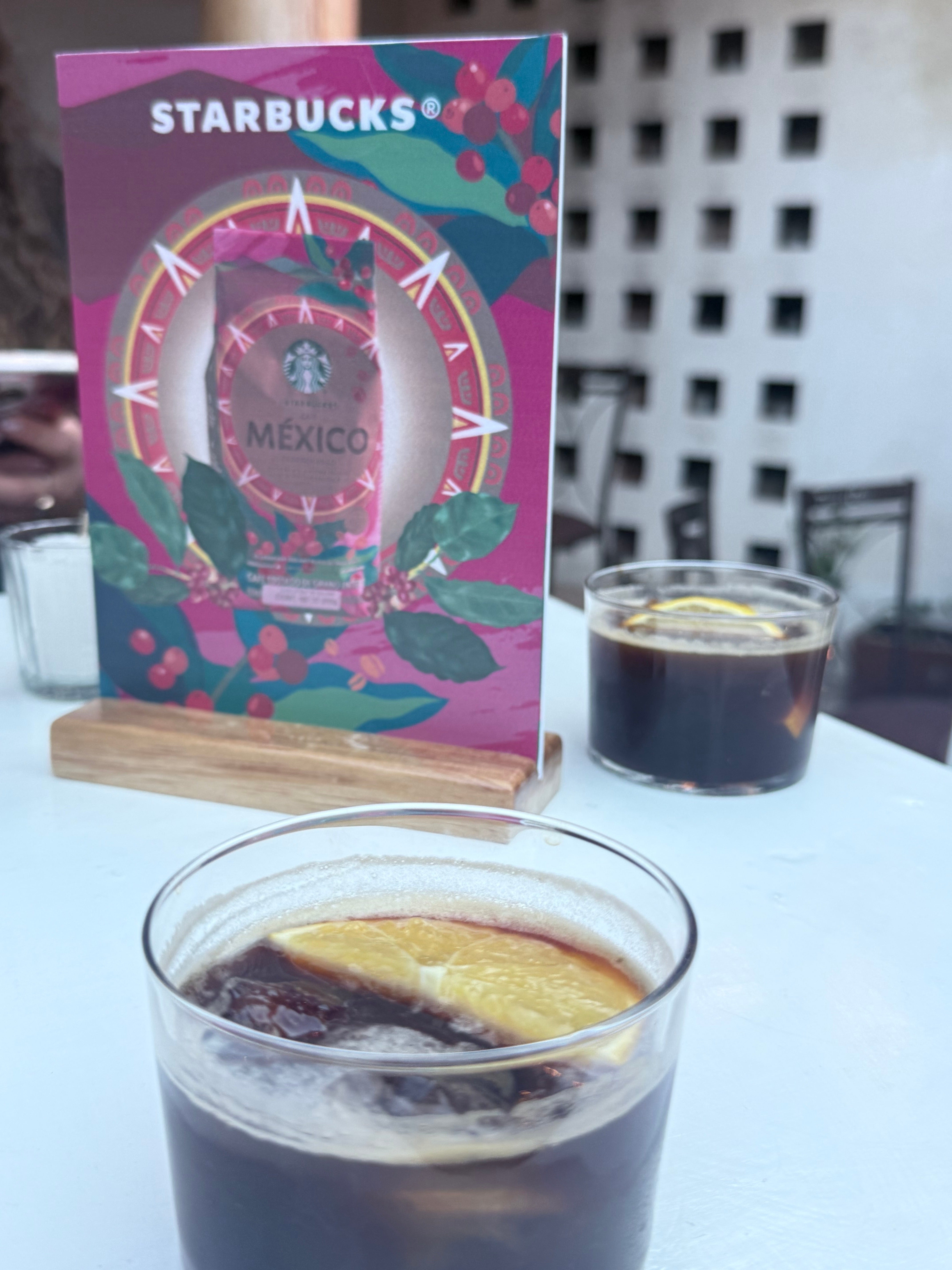 Vasos de café frío con rodajas de naranja frente a un cartel de Starbucks que dice "México" en una mesa blanca