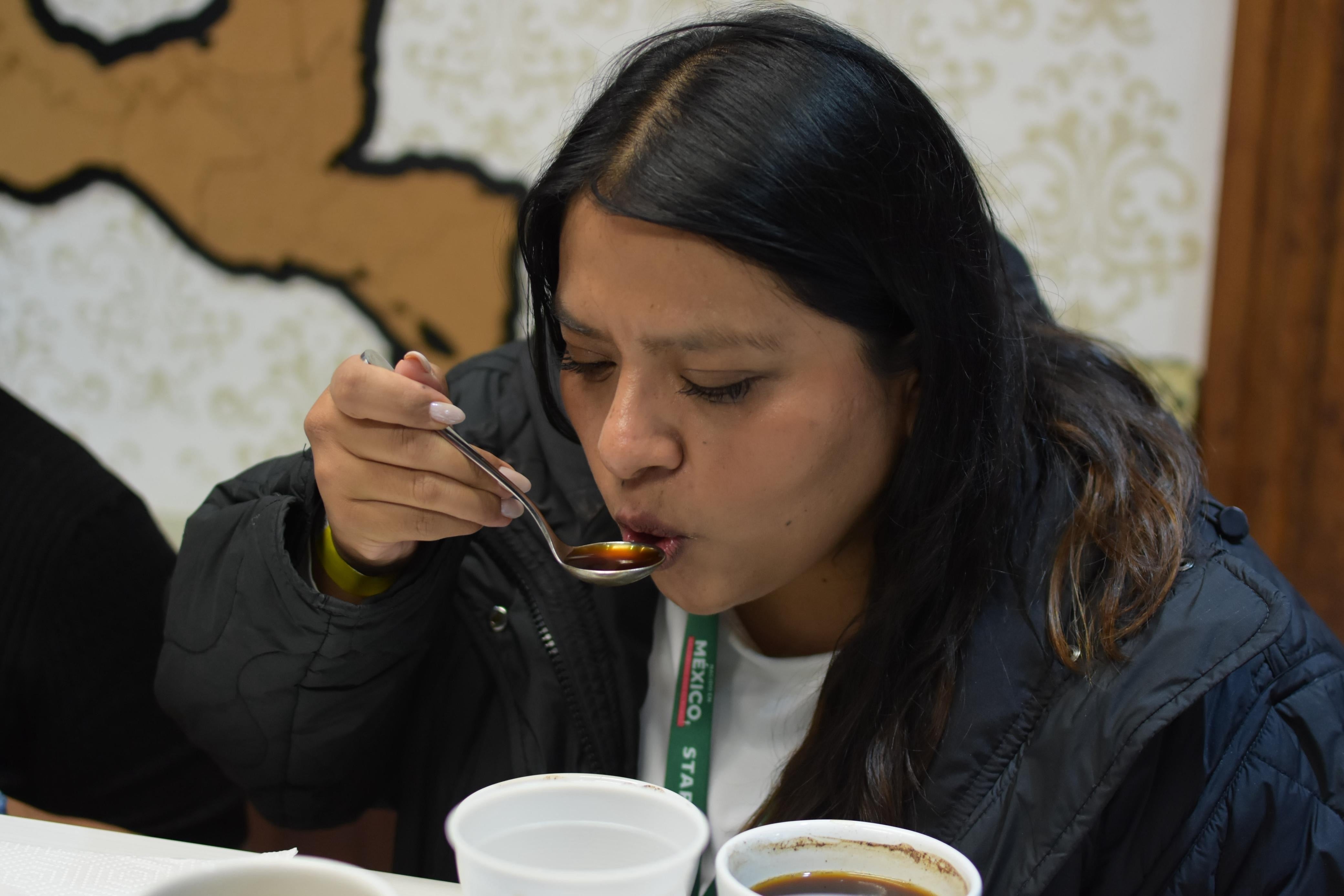 Persona probando una bebida con una cuchara en un evento. Concentrada en el sabor, con tazas y un fondo decorativo visible
