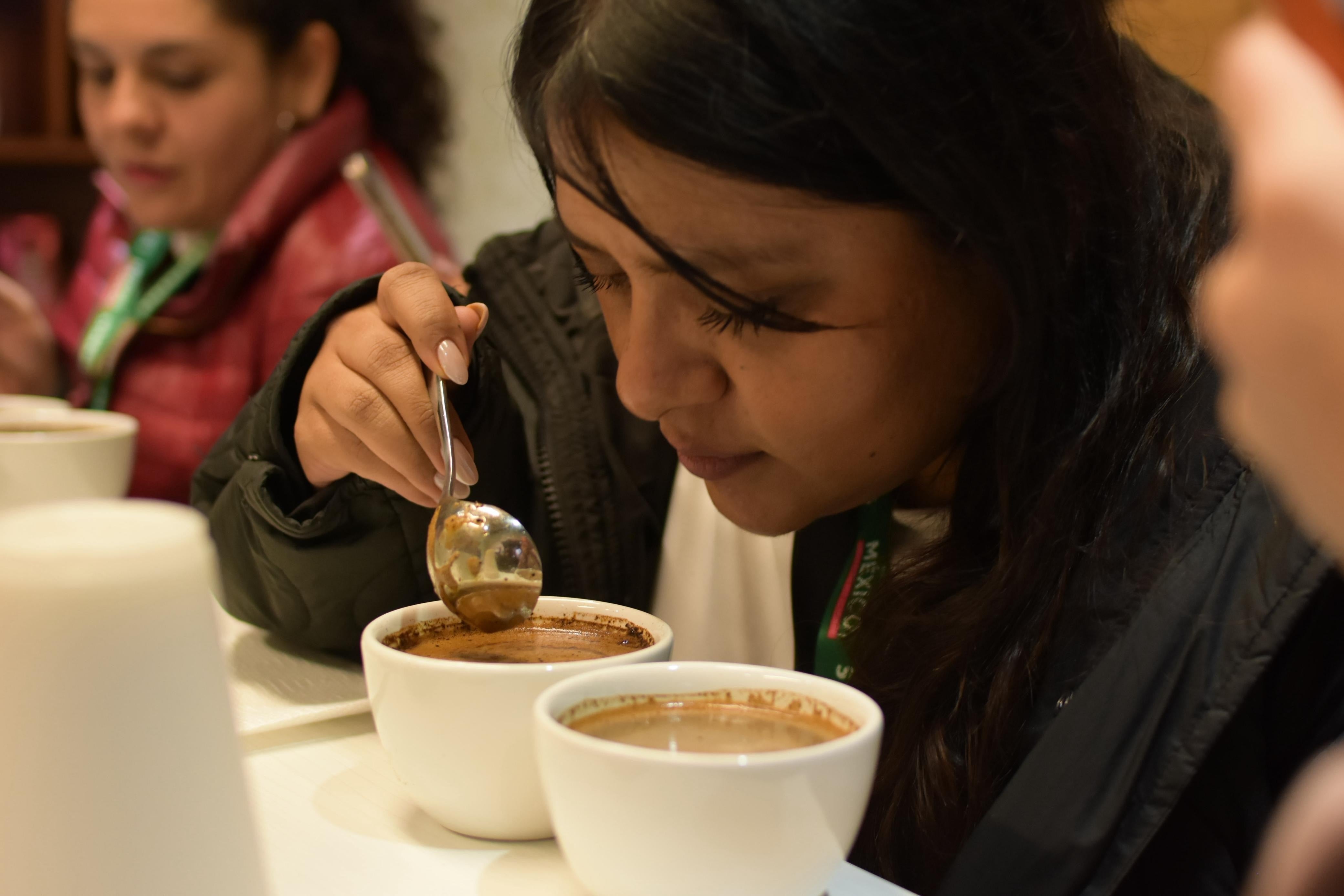 Persona probando una taza de café, evaluando el aroma. Al fondo, otra persona también tiene una taza. Ambiente de degustación