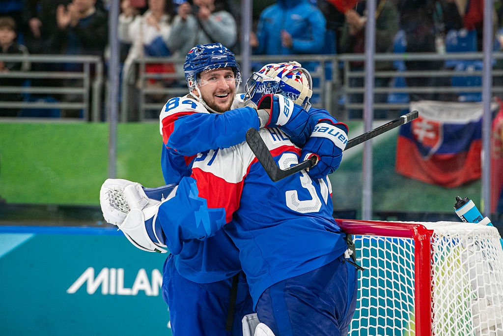 Hockeykeeper og lagkamerat feirer seier på is; de omfavner med glede nær målstolpen, iført blå uniformer med verneutstyr