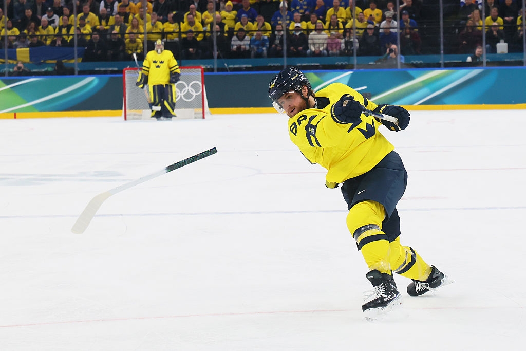 Hockeyspiller i aksjon, skyter pucken på is under en kamp. Lagmedlemmer og målvakt synlig i bakgrunnen