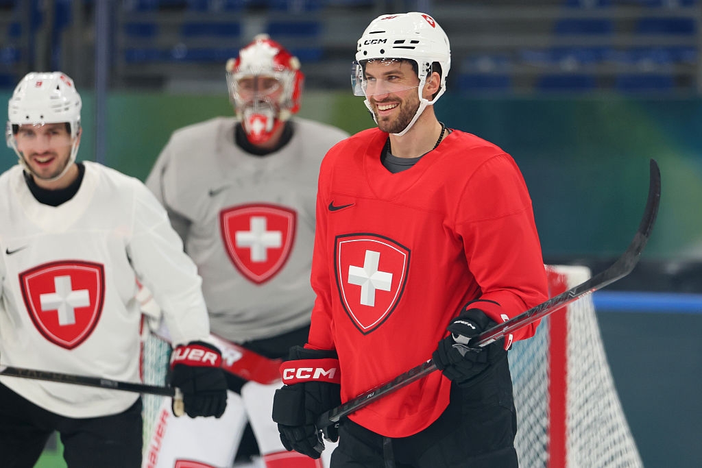 Hockeyspillere i lagdrakter trener på is; en i rød trikot, andre i hvit. De smiler og holder hockeykøller i nærheten av nettet