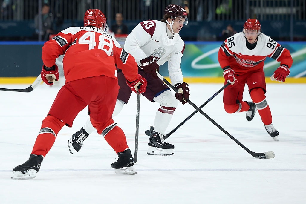 Ishockeyspillere i aksjon under en kamp; to bruker rødt og en i hvitt fokuserer på pucken. Arenasetting med tilskuere i bakgrunnen