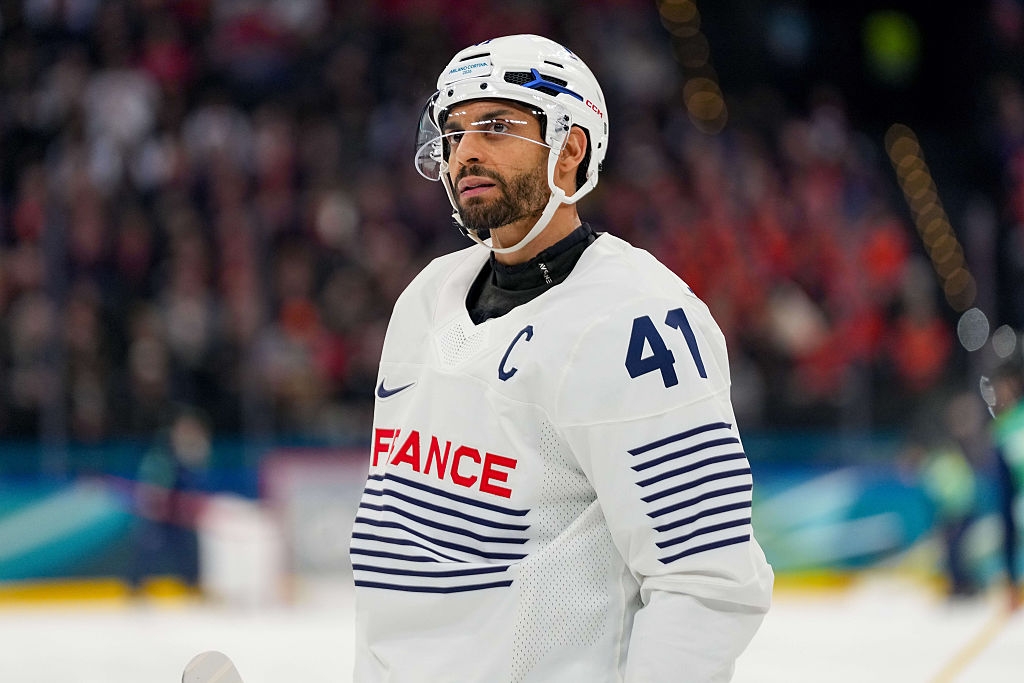 Hockeyspiller i hvit trøye og hjelm står på isen med "Frankrike" på forsiden, ser mot siden