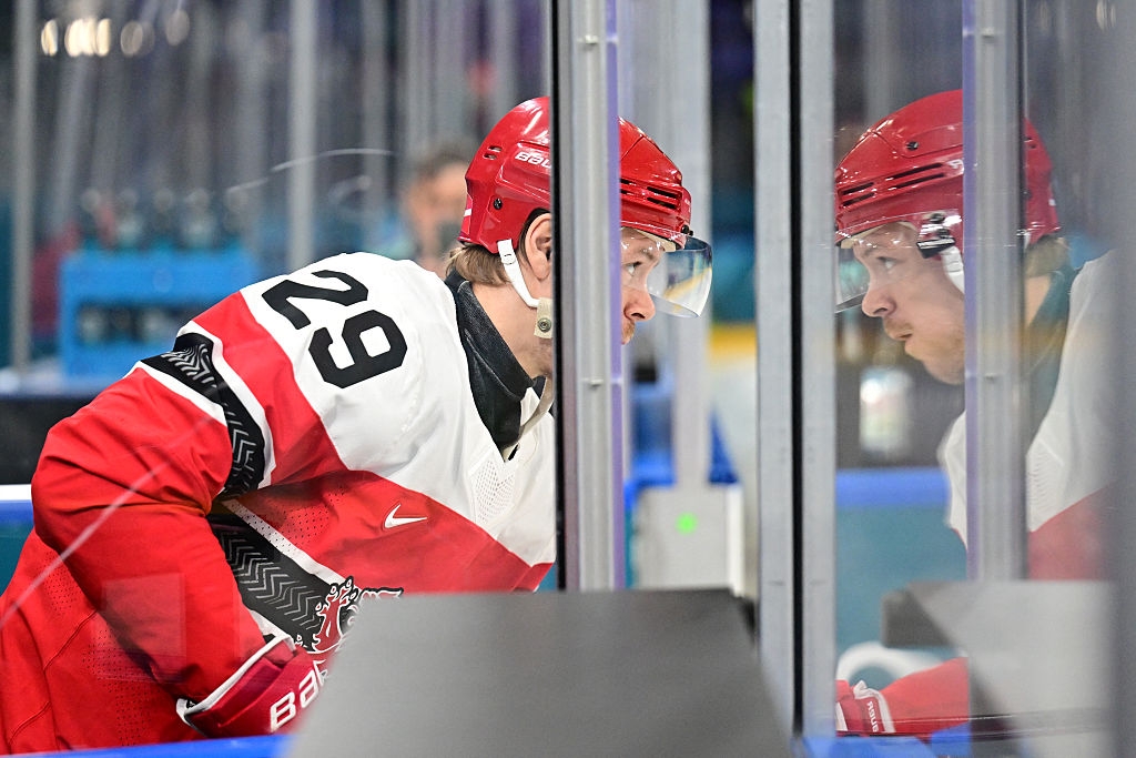 Hockeyspiller iført rød og hvit uniform fokuserer intenst, vendt mot et reflekterende glasspanel på en kamp