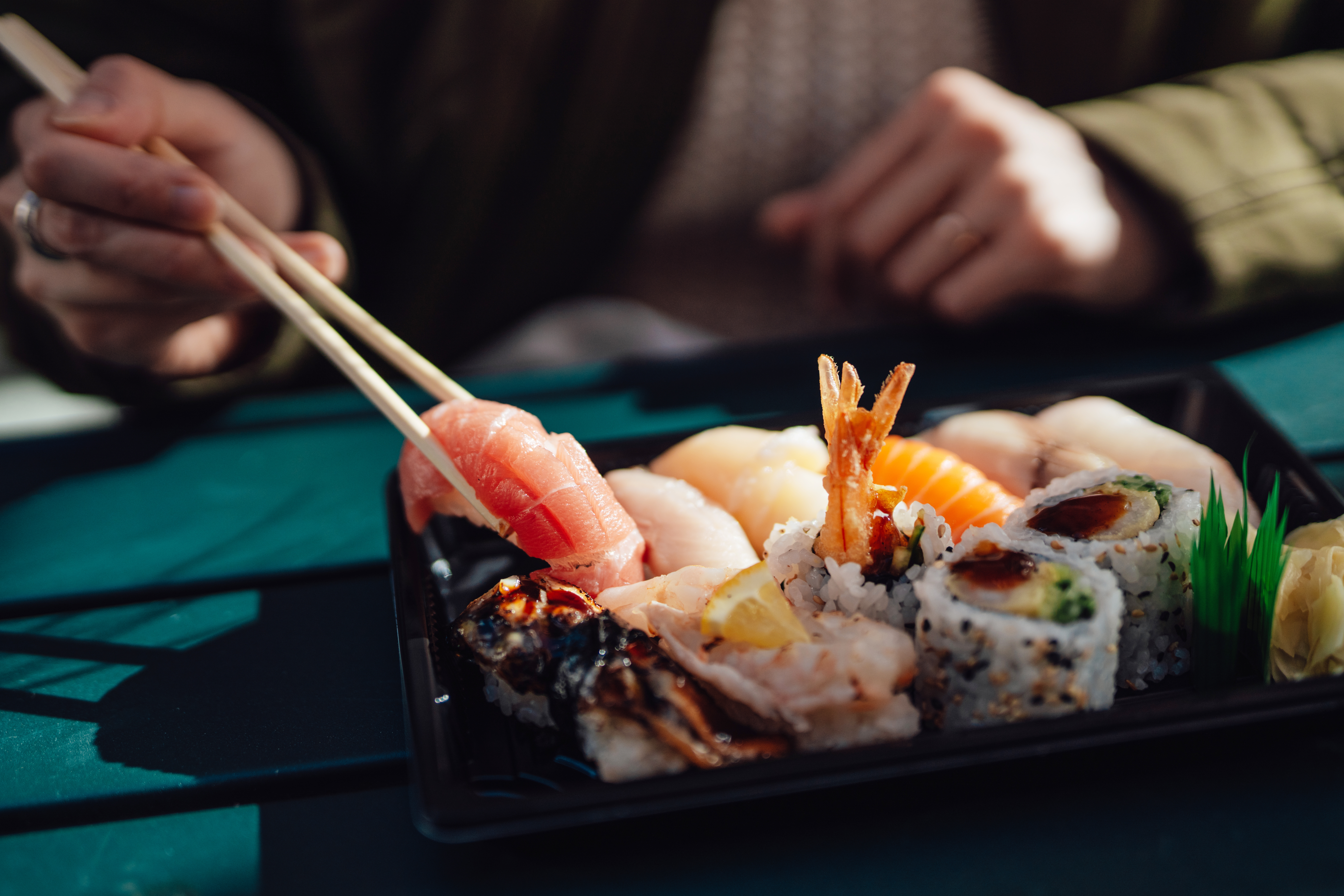 A person uses chopsticks to pick up a piece of sushi from a tray, featuring various sushi and rolls on a table