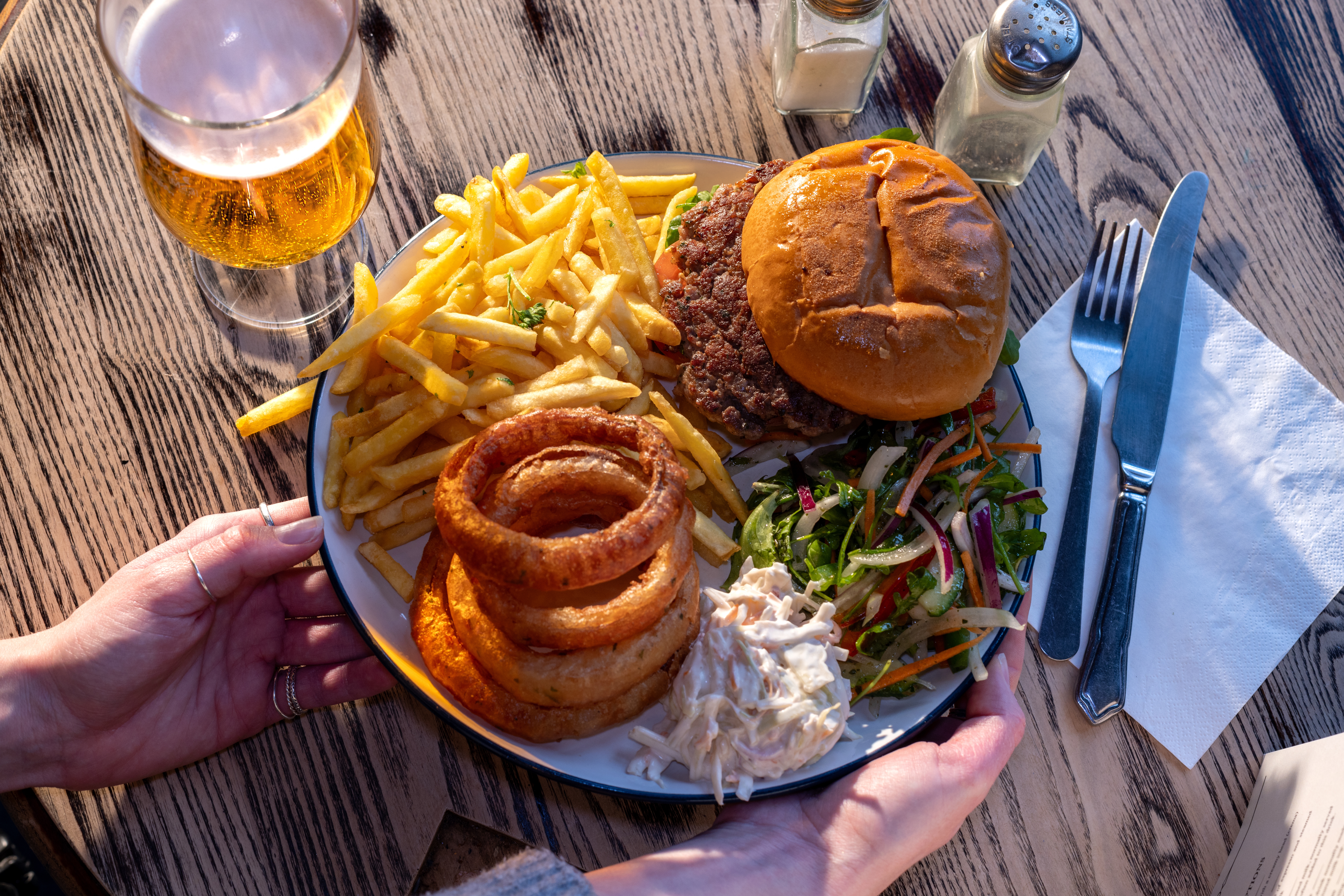 Burger with fries, onion rings, coleslaw, and salad on a plate, placed on a wooden table with a glass of beer and cutlery. Hands adjusting the plate