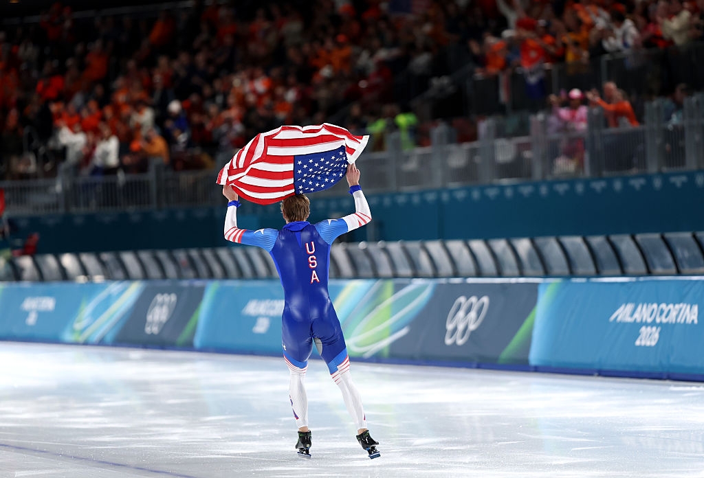 An jock successful a USA speedskating azygous holds an American emblem connected an Olympic crystal rink, celebrating successful beforehand of a cheering crowd