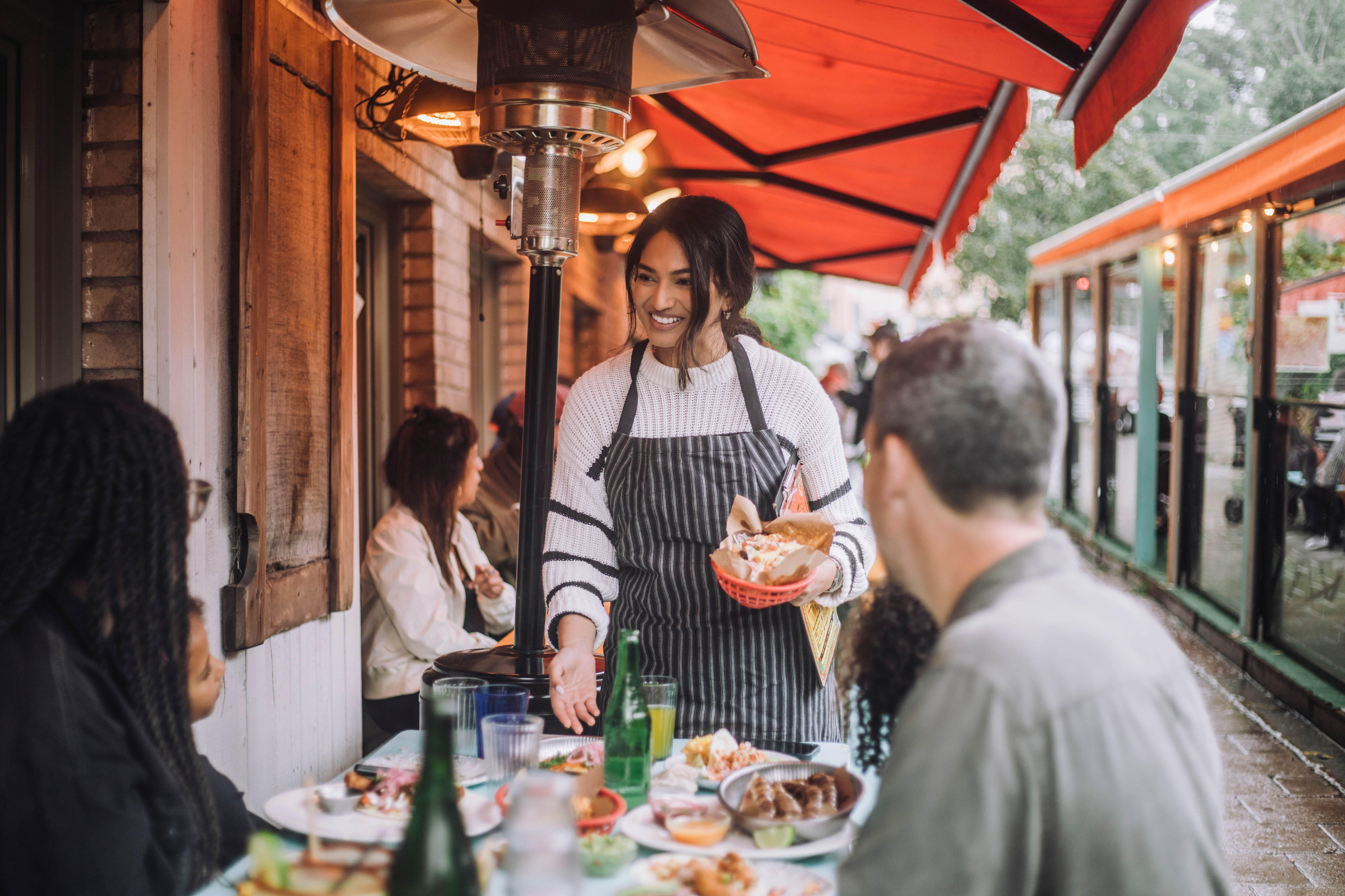 Person smiling and serving a meal at an outdoor table, engaged in lively conversation with diners in a cozy restaurant setting