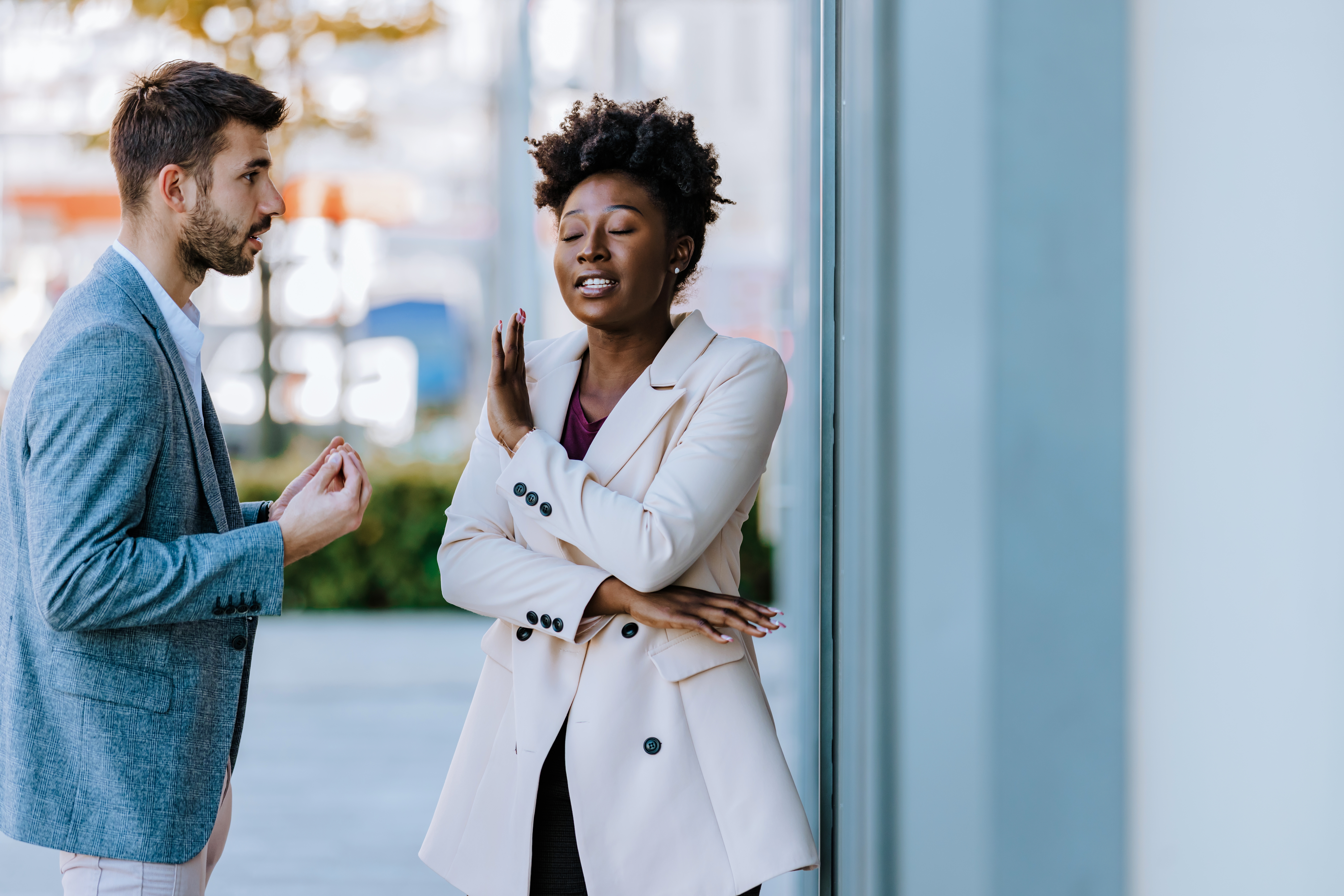 A woman in a stylish blazer raises her hand while speaking to a man wearing a suit, both engaged in a serious conversation outdoors