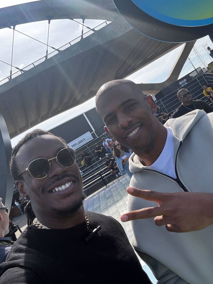 Two individuals airs  outdoors astatine  an event. One wears sunglasses, and the different   shows a bid    sign. People and a modern   operation   are successful  the background