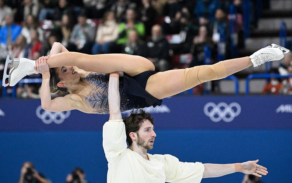 Figure skating duo performs a lift at an ice rink during a competition, with an audience watching in the background
