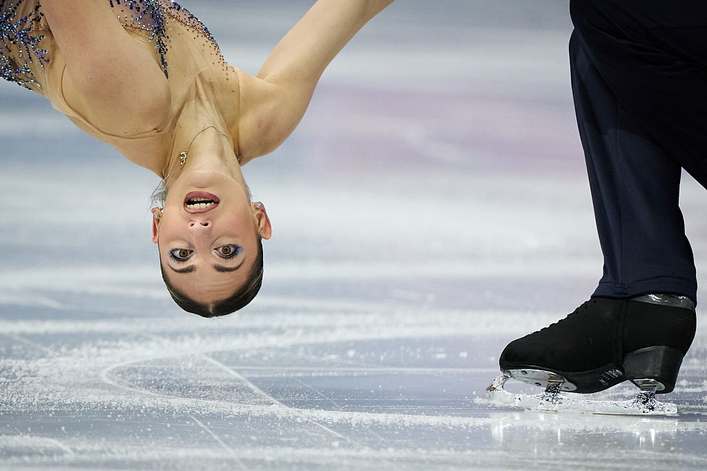 Kunstløper i en elegant positur på is, midt i ytelse, med fokus på skaterens ansikt og partnerens fot i bevegelse
