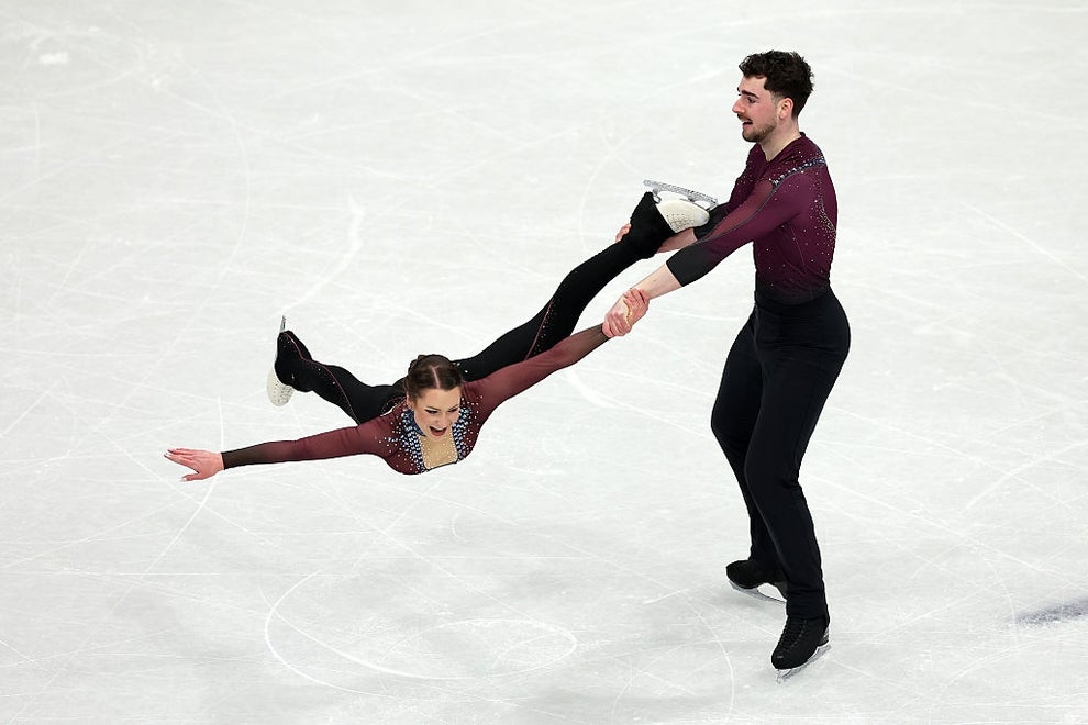 Two ice skaters perform a lift, with the man holding the woman horizontally by one arm and leg during a routine on the ice rink